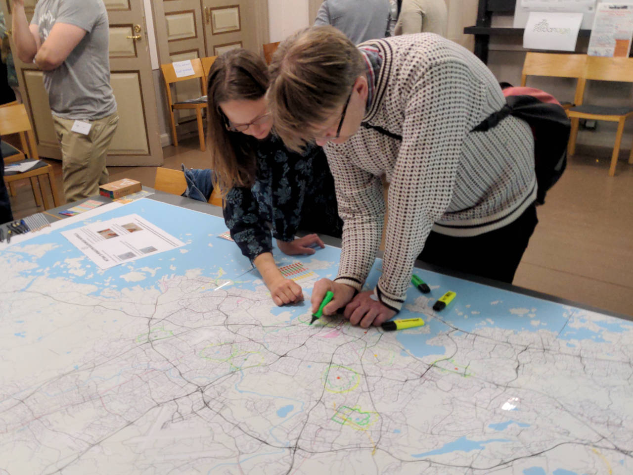 Two visitors lean over a large map (1.5 x 3.5 metres) of the Helsinki metropolitan area, drawing the area covered by 15 minutes walking around an important location to their lives