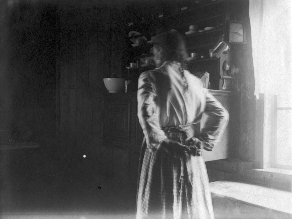 Mustavalkokuvassa nainen seisoo vanhanaikaisessa pirtissä selin kameraan. Hän sitoo lankaa kädet selkänsä takana.