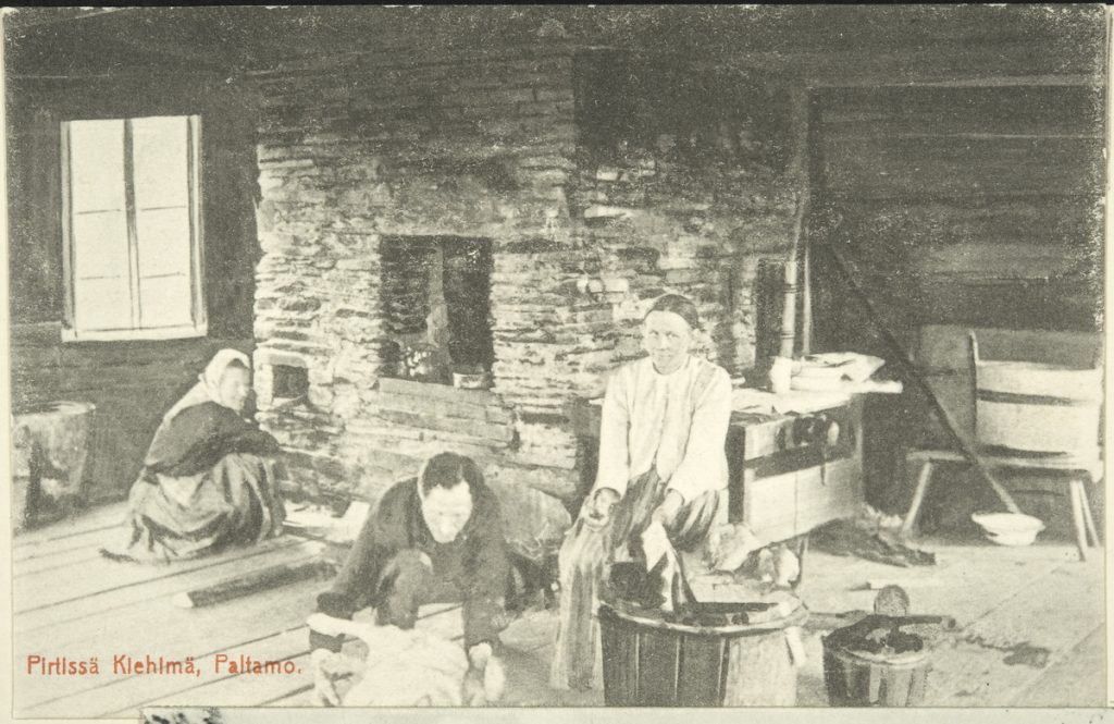 Kuvassa kolme henkilöä istuu vanhassa pirtissä suuren leivinuunin tai takan edessä.