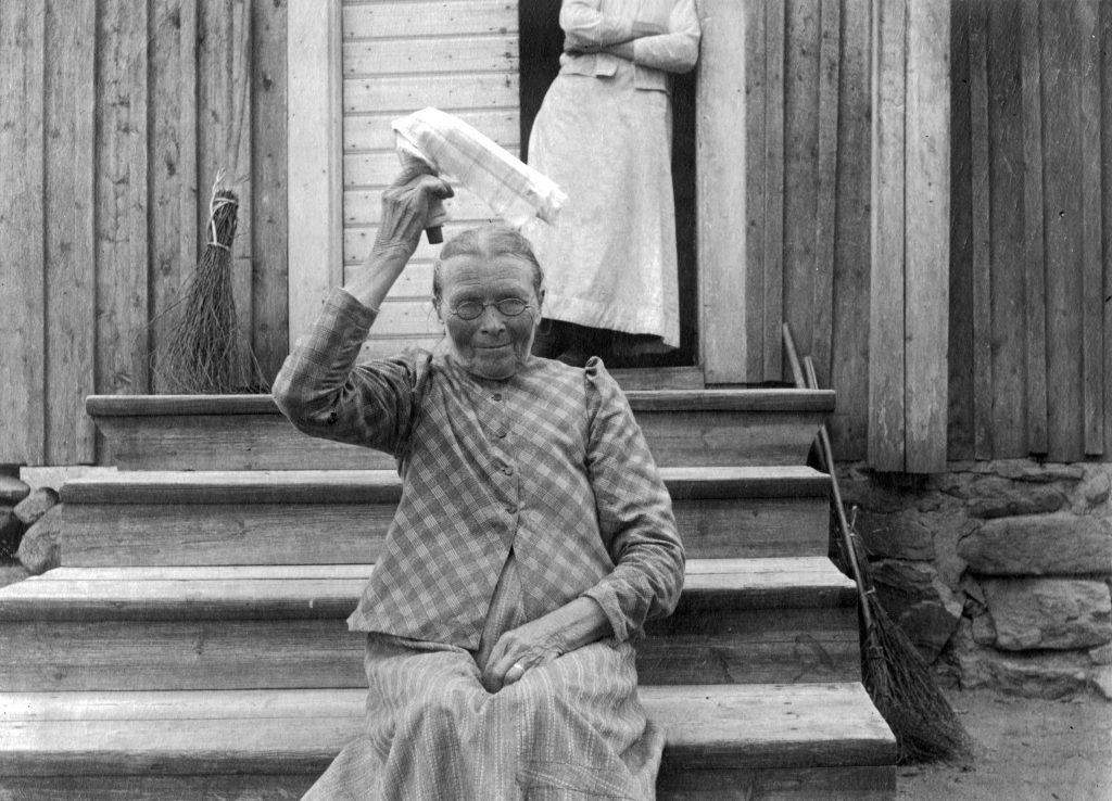 Vanha nainen istuu puutalon ulkoportaalla ja heiluttaa päänsä päällä kangaspalaa.