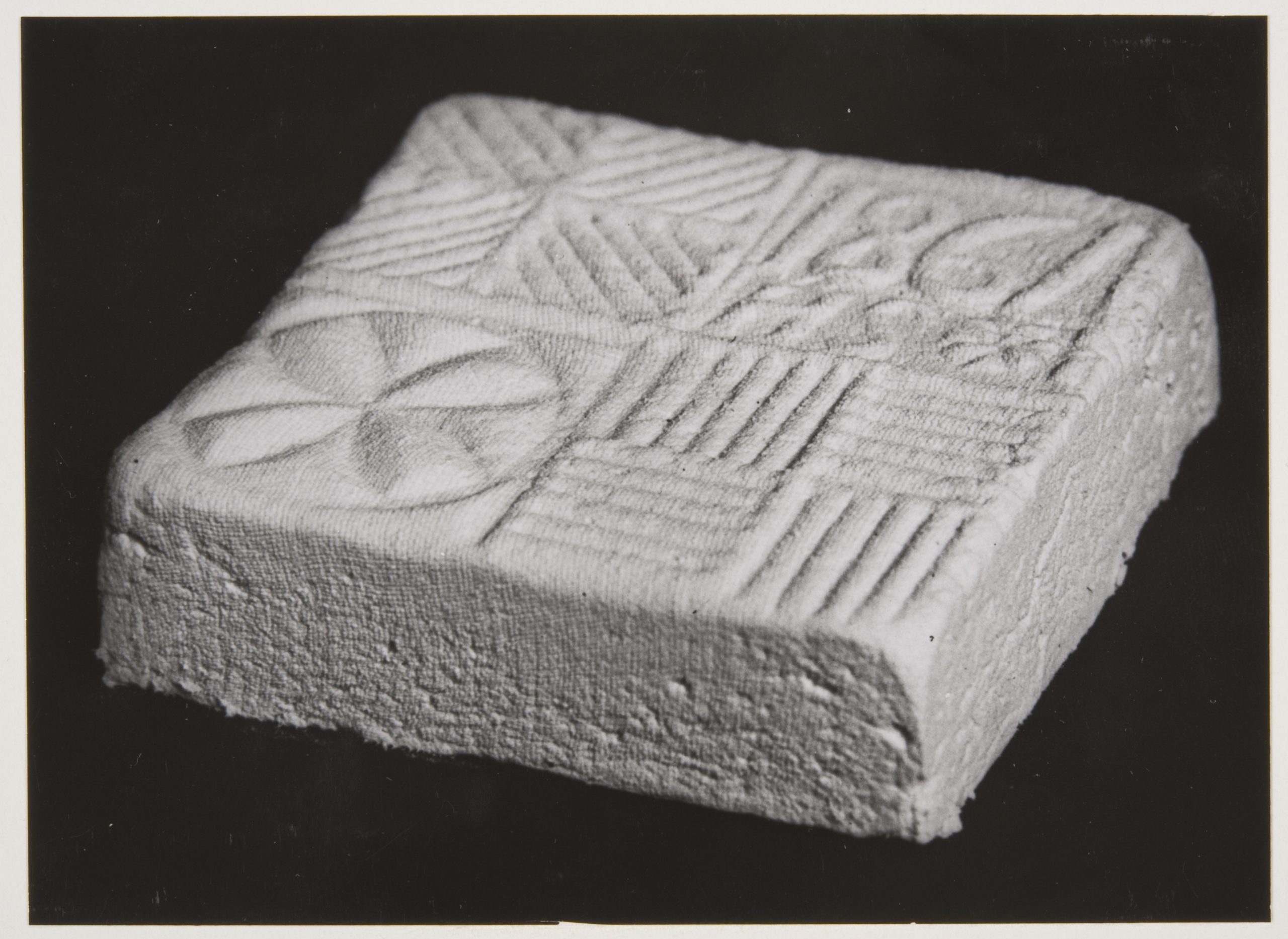 Svartvitt fotografi av en ost som beretts i ostform. Osten är låg och fyrkantig. En dekoration från formens botten syns på ostens ovansida.