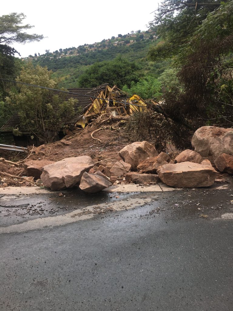 A road is in the foreground with a green hill in the background. There is a gash of dirt and giant rocks strewn across the road where there has been a landslide.