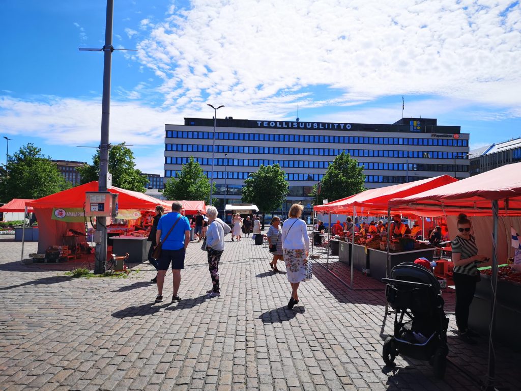 There are several people walking through and standing in an outdoor food market. The sky is blue with white clouds. 