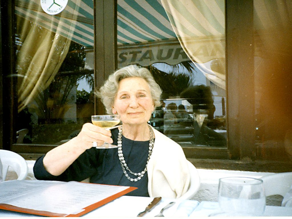 En kvinna med grått hår sitter vid borden vinglas i handen.