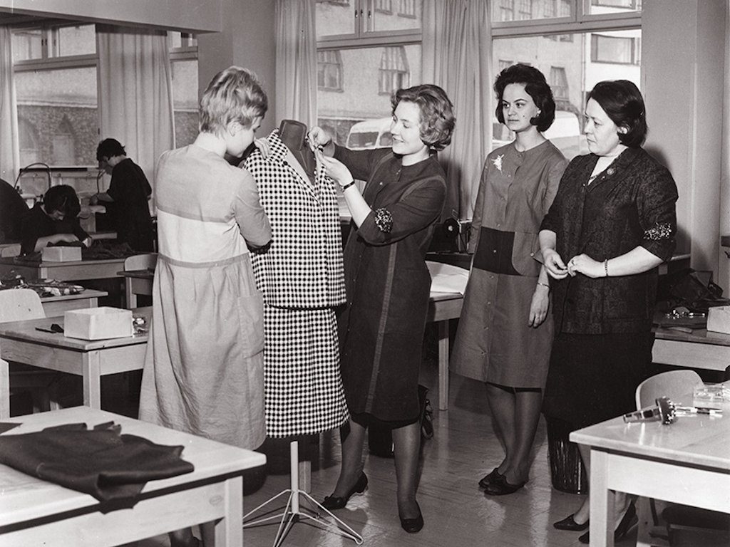 Ett svart-vitt foto av fyra damer som tittar på en färdig klänning på en torso i klassrummet.