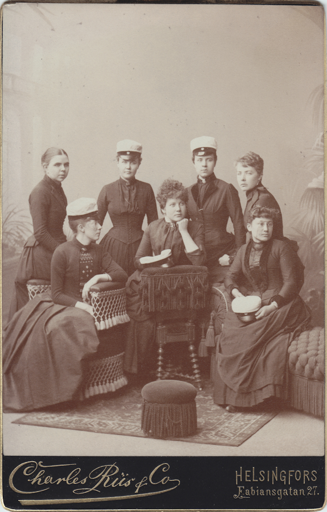 Sju unga kvinnor, varav fyra står i bakre raden. De två i mitten bär studentmössa på huvudet. I främre raden sitter tre kvinnor, varav den till vänster har vänt sig i sidled och bär studentmössa på huvudet. De övriga två håller sina mössor i famnen.