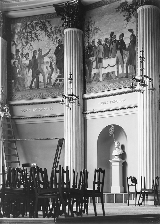 Ett gammalt fotografi som visar de ursprungliga Florafesten-målningarna i universitetets festsal. Målningarna sitter högt uppe på väggen. I förgrunden syns högar med staplade stolar.