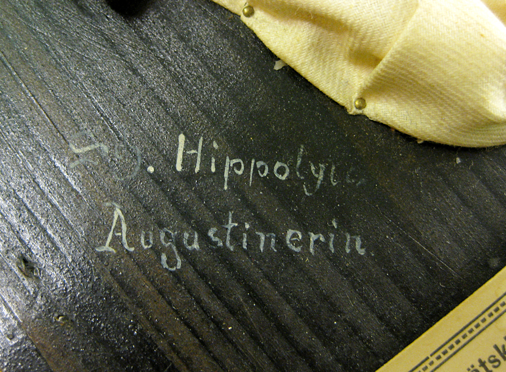 Närbild av en signering som målats med vit färg på svartmålat trä. Litet vitt tyg och två nålar som håller tyget på plats skymtar uppe i det högra hörnet. En del av vaxavgjutningens etikett skymtar nedtill i det högra hörnet.