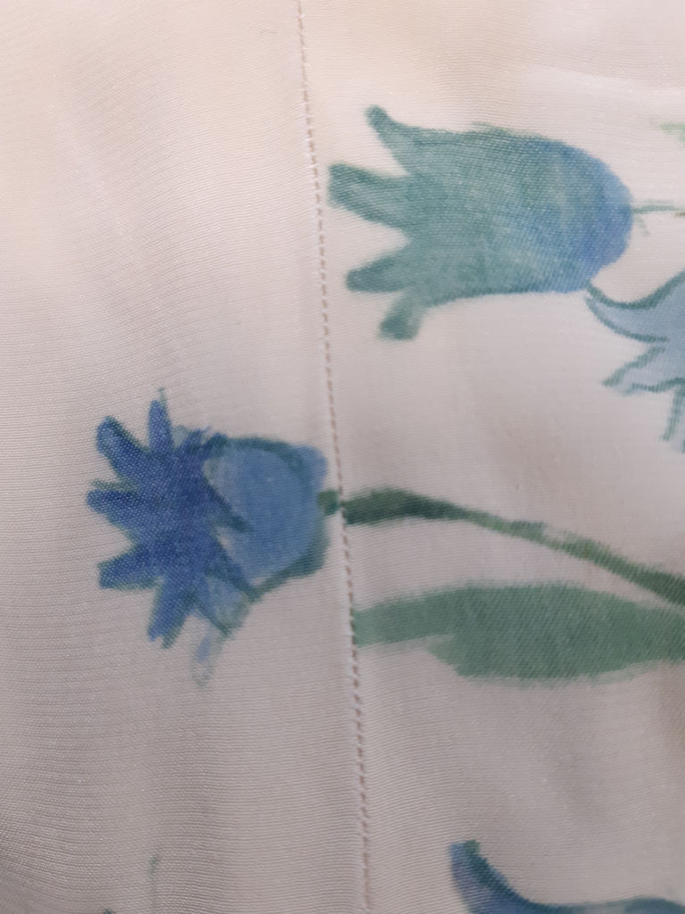 En blå klockformad blomma målad på ett vitt tyg. Blommans stjälk bryter vid sömmen.