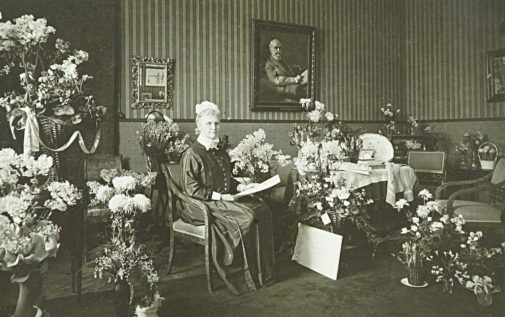 En dam som är klädd i mörk klänning och vit hätta sitter i en fåtölj bredvid ett bord. På bordet, på golvet omkring damen och på sidobord har ställts många blomsterarrangemang. 