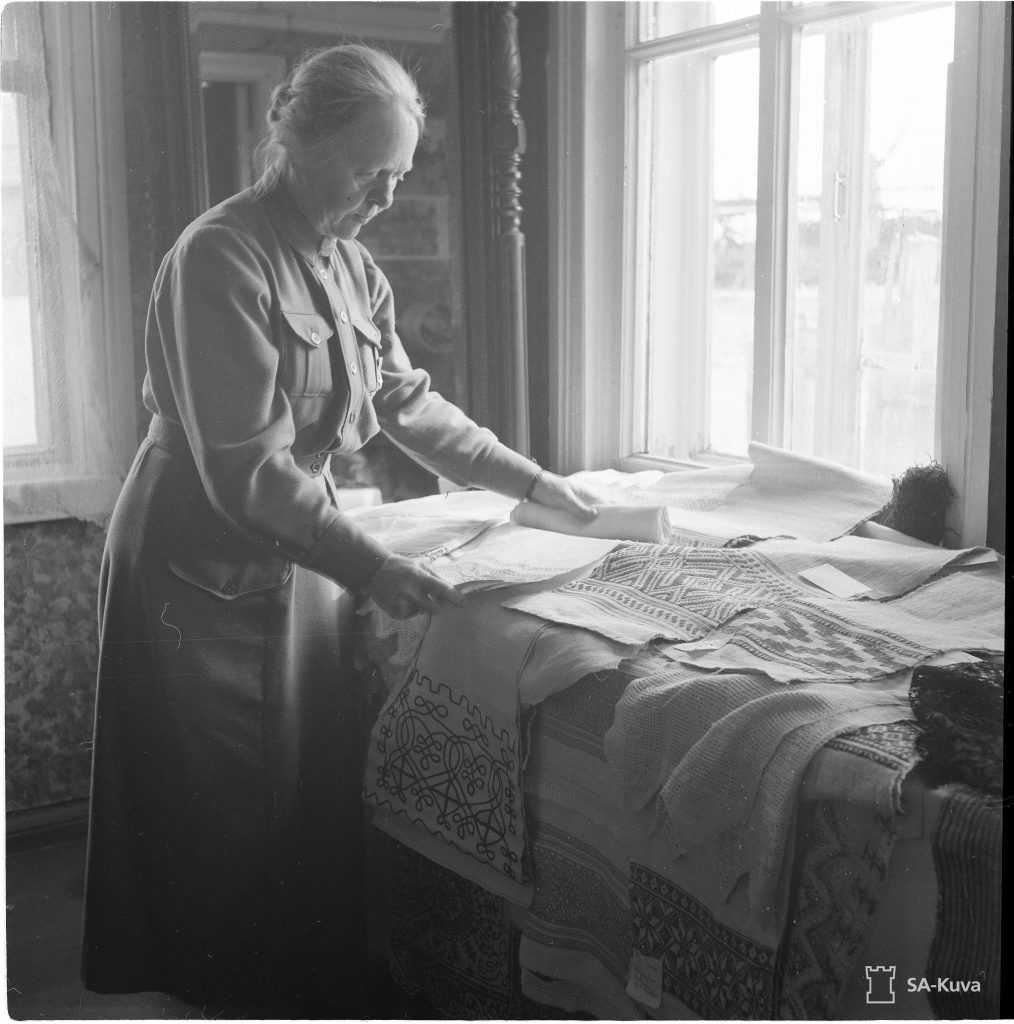 En äldre dam klädd i lottauniform står vid ett bord vid ett fönster. Hon studerar textilier som ligger på bordet.