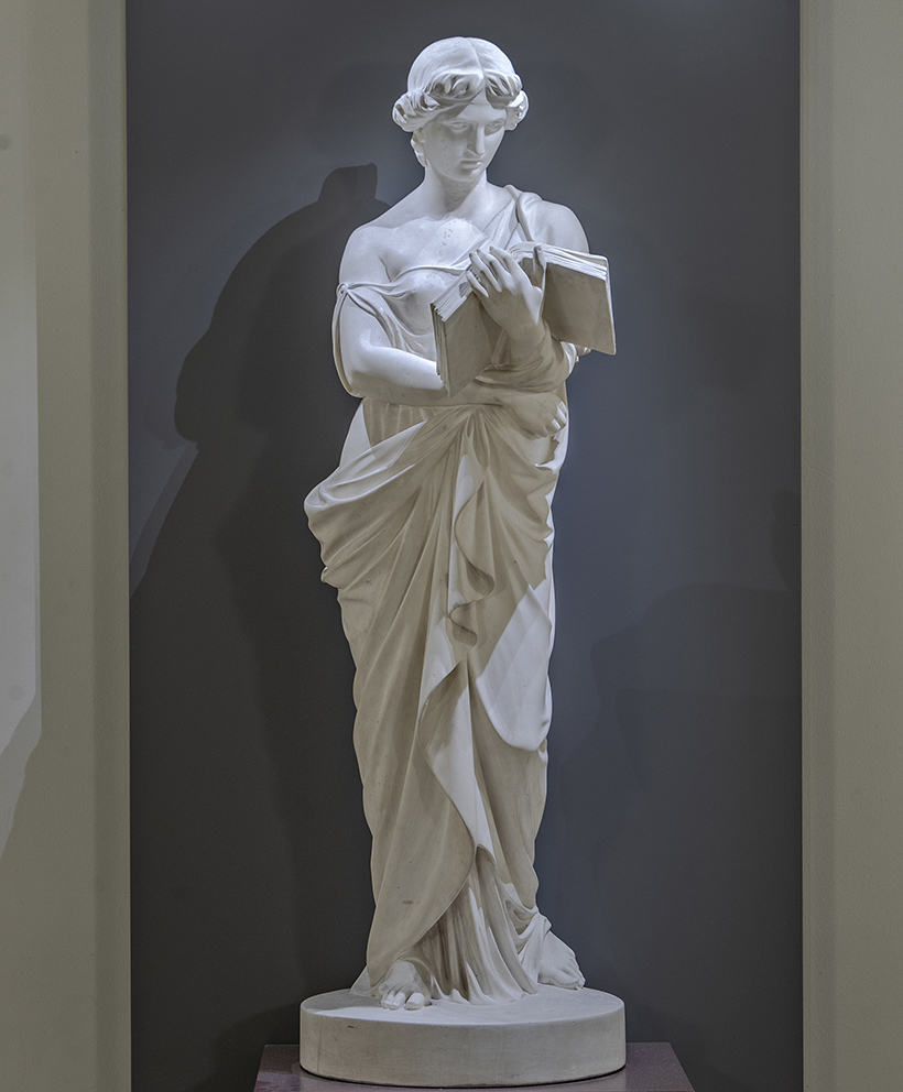 En vit skulptur föreställande en kvinna, på en rund, låg piedestal.