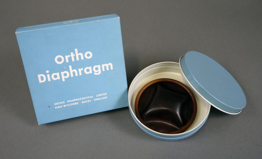 Ett runt och platt gummiföremål i en rund ljusblå metallask. Bredvid asken står en ljusblå kartongförpackning med texten Ortho Diaphragm.