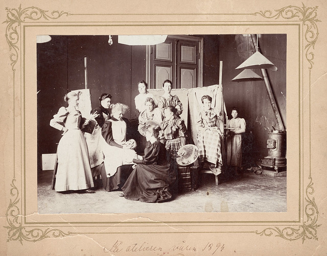 A black and white photograph of 10 women, standing and sitting. dressed with long skirts. In the background two cloths, a long door and a stove.