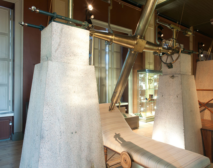 Two large stone slabs supporting an old telescope. Beneath the telescope is an upholstered bed-like piece of furniture with one end elevated higher than the other.