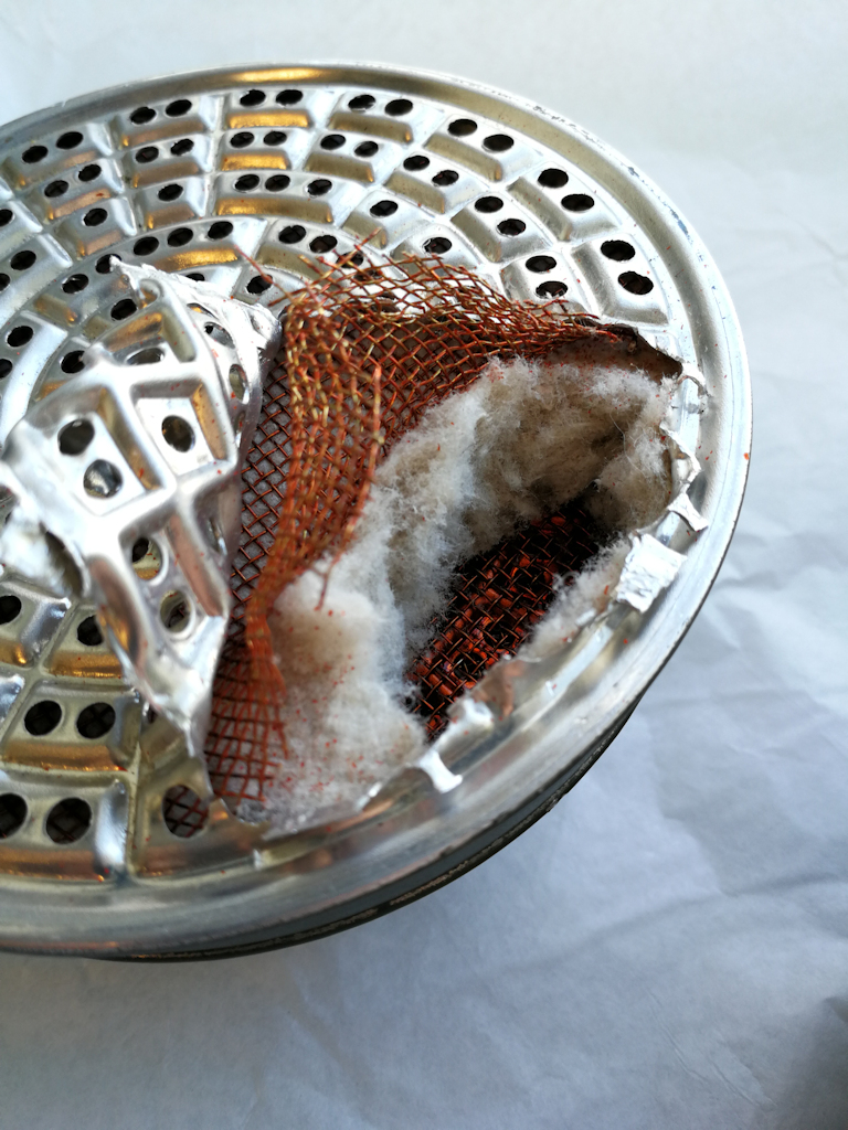 The partially opened filter cartridge of a civil defence gas mask. Under the mesh metal plate, denser metal netting can be seen, beneath which is a layer of white cotton-type filter material. 