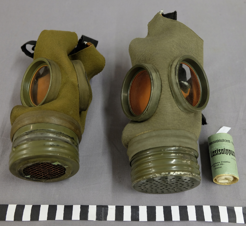 Two gas masks side by side. The masks are made of rubber covered by a green fabric. Below the masks is a scale. Next to one of the masks is a small cylinder-shaped package with the word Lasisaippua (‘Glass soap’).