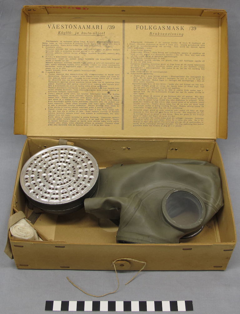 Green gas mask in a cardboard box. Finnish- and Swedish-language instructions for the use and care of the mask are printed inside the lid of the cardboard box.