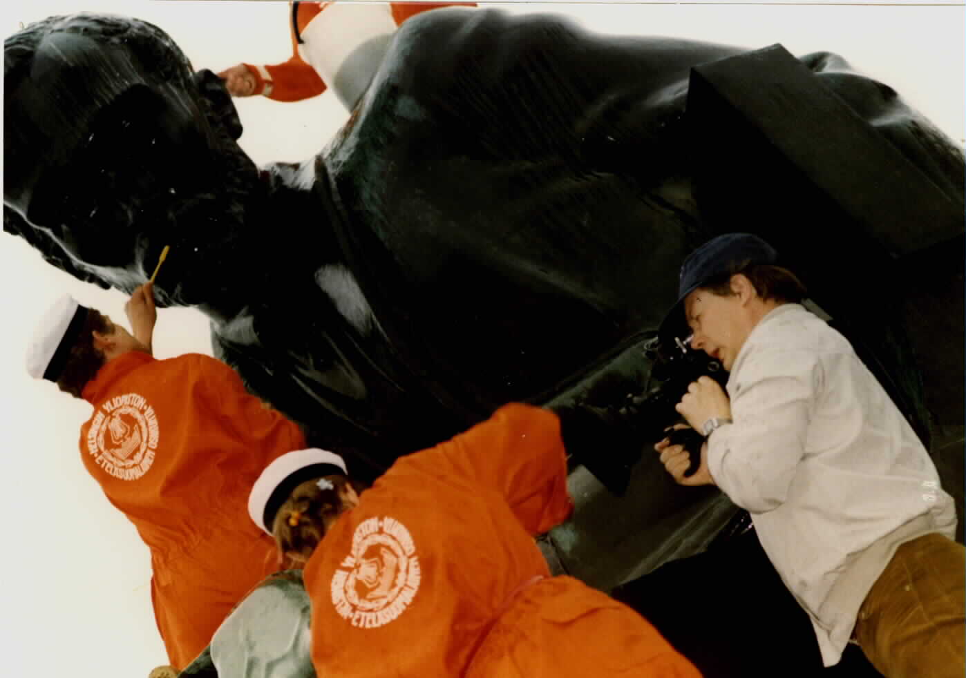 Two students, with their backs to the photographer, wearing orange overalls and student caps while washing the statue of writer Aleksis Kivi with toothbrushes. Another student on the right is recording the event.