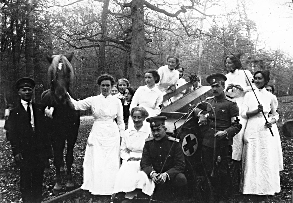 A photograph taken outdoors showing nurses in white outfits and doctors in uniforms, possibly on an excursion. The group poses next to a horse and cart featuring the Red Cross emblem. Trees can be seen in the background. 