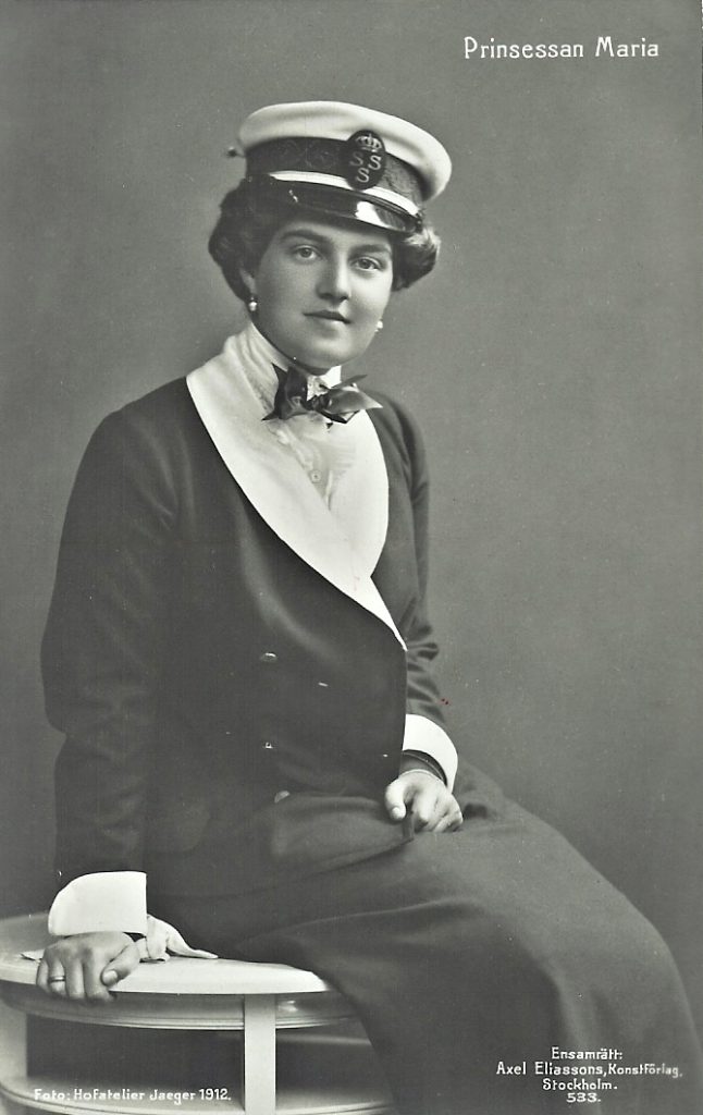 A woman wearing a dark jacket and skirt, a white shirt and a peaked cap is sitting on a bench and looking straight into the camera.