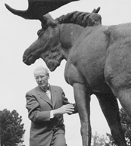 A black-and-white photo taken from the back left-hand side of a bull elk sculpture, next to which stands an elderly man wearing glasses, a suit, a dress shirt and a bow tie.