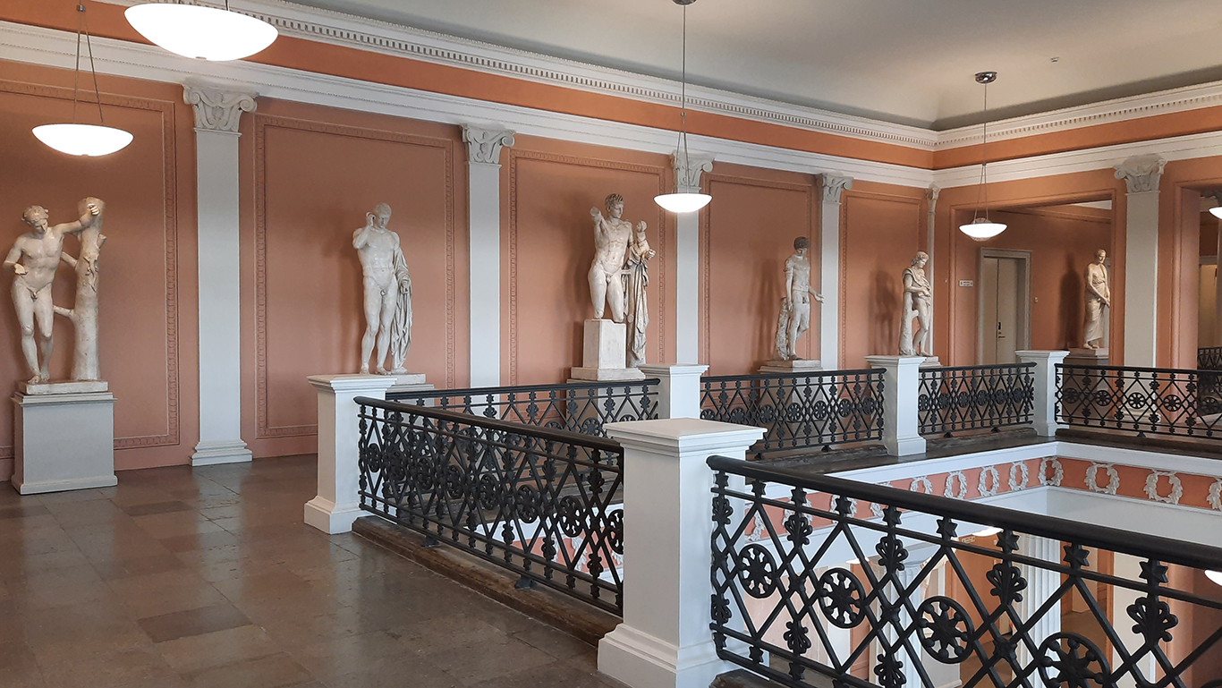 A view from the fourth floor of the University’s Main Building. On the right is the vestibule behind a wrought-iron banister, and on the back wall is a row of sculptures in a corridor.