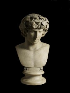 A photo of a marble bust against a black background, showing a young man with a bare upper body, curly hair and a wreath on his head.