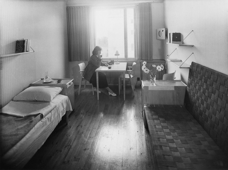 A woman sitting by a desk next to a window, with a bed at front left and a sofa on the right.