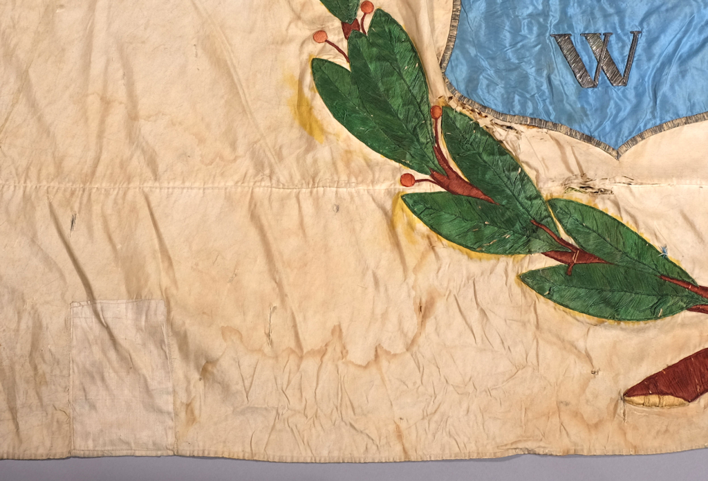 The photo shows damage to the lower corner of the flag: there are yellow stains next to the leaves embroidered with green thread. There are tears above the leaves and at the bottom there are moisture streaks, i.e. light brown wavy patterns of dirt accumulated by moisture.