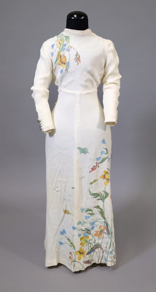 A white evening dress with a multi-coloured pattern similar to a flower meadow on its hem and front. The dress has a close-fitting top, long sleeves and a high neckline that covers some of the neck. The dress is long, has a short train and flares at the hem.