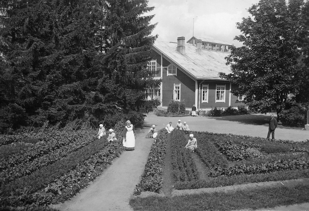 Two vegetable plots intersected by a narrow gravel road. Seven women are squatting among the plants, one with a dog under her arm. A woman in a white apron and cap is standing on the road. A man in a suit is standing behind the vegetable plot on the right. Behind the plots are a wider gravel road and trees, through which a two-story building can be seen. A woman wearing a hat or scarf is standing in front of the building. The sun is shining, and one of the windows of the building is open.