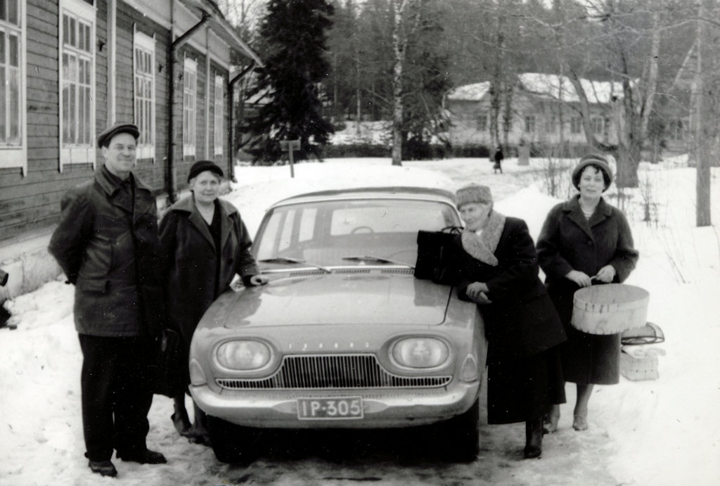 A winter landscape with wooden buildings in the background. In the centre of the photo is a car surrounded by four people. 