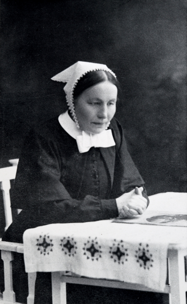 A sombre woman sits at a table with her hands clasped. She is wearing a white bonnet and a dark dress.