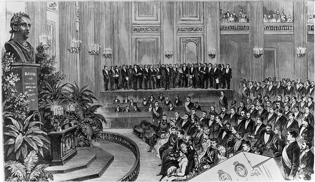 A black-and-white drawing of the University’s Great Hall. Men and women in festive attire are seated, while a choir is performing on stage in the background. On the left is the large sculpture of the upper body of a man on a high stand, surrounded by several plants. In front of the sculpture is an ornate podium, on which two candelabra are standing.