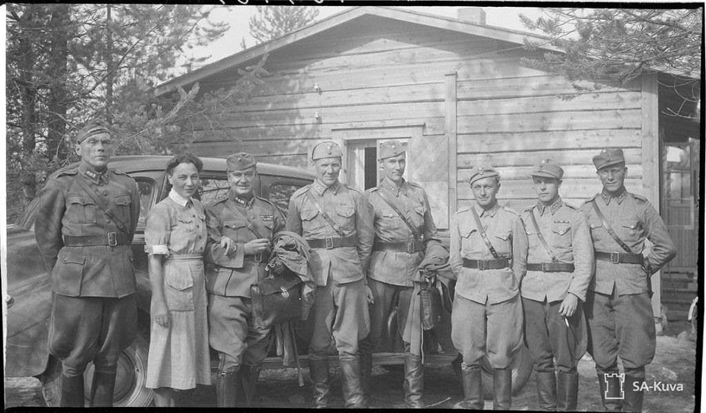 Seven soldiers and one member of the voluntary paramilitary Lotta Svärd organisation in a group photo in front of a car outside a log building.