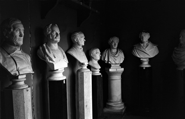 A black-and-white photo of six white busts of short-haired men in a semicircle. Some of the men have beards and sideburns. The statues are placed on high, narrow pedestals.