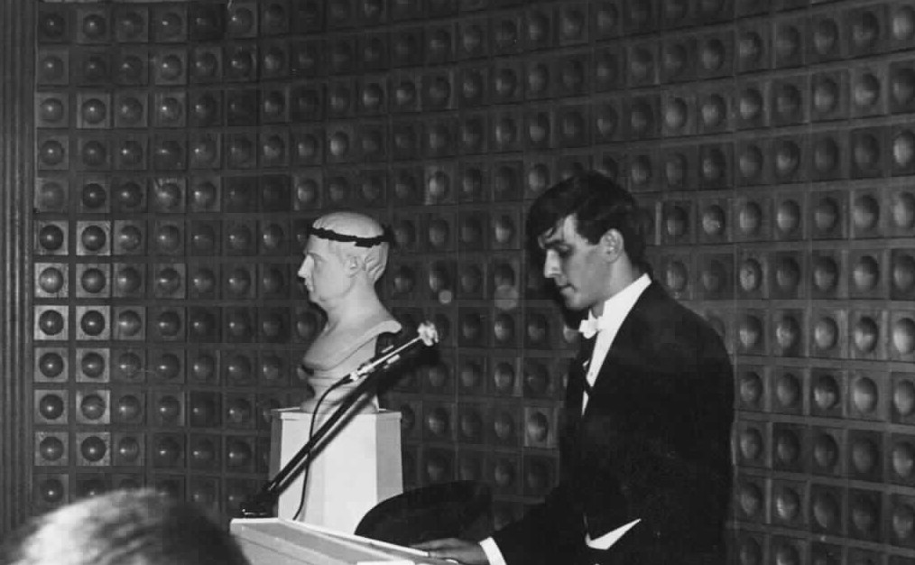 A dark-haired, tailcoated young man seen from the side stands at a podium with a microphone on it. Next to him is a white sculpture of a man with a wreath placed on the head. In the background is a wall with spherical recesses in rows.