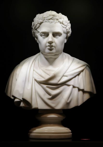 A white marble bust of a short-haired man wearing a wreath and a robe, of which only the top part is visible. The bust is placed against a black background. The statue has a low, round base.