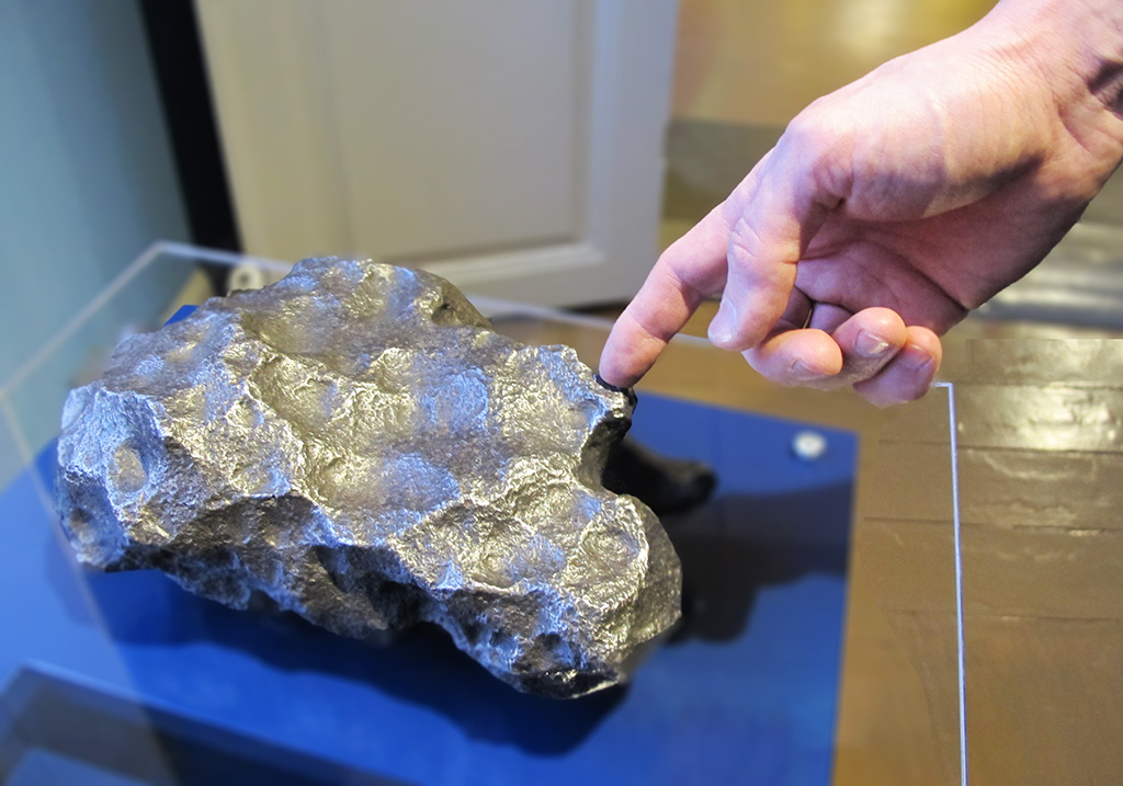 A shiny metallic meteorite resting on a blue table. A hand emerging from the top right corner of the photo touches the meteorite with one finger.