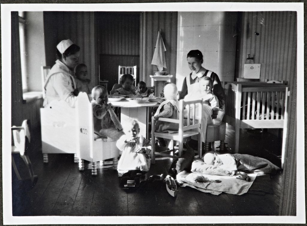 Two nurses and eight children at a round table set with soup plates. 