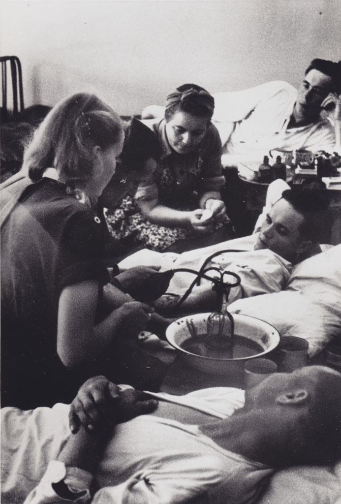 Three male patients lie in hospital beds. Two women and a man sit either side of the central bed. The man sitting is giving one of the patients a transfusion from a glass bottle placed in a wash basin half full of water. The blood has coloured the water red. All six people in the picture are following the transfusion procedure closely.