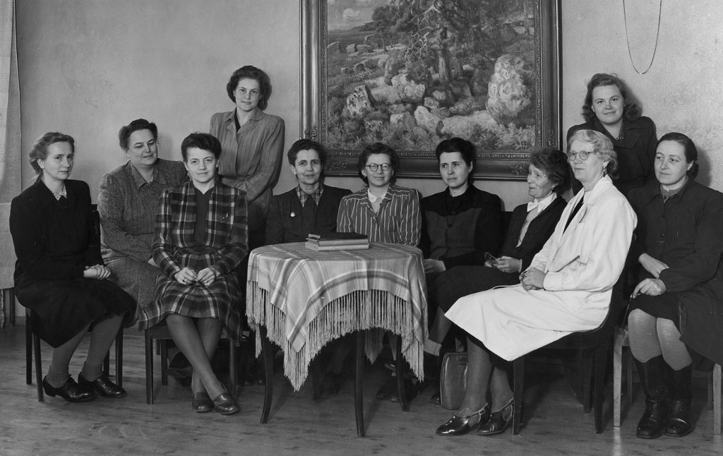 Eleven women in skirt suits are sitting on a sofa and chairs at a small table. Above the sofa on the wall is Helmi Biese’s painting Vanha mänty (‘An Old Pine’) depicting a pine tree growing on a rocky bank.