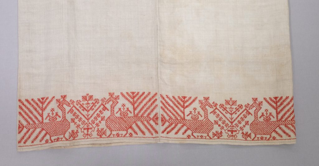 The hem of a white garment embroidered with two pairs of birds facing each other, their tails forming geometric shapes.