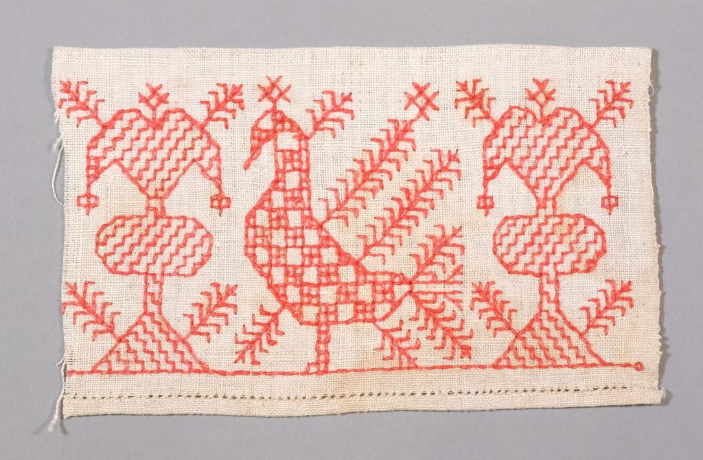 A rectangular piece of cloth with a stylised tree embroidered in red at both ends and, in the centre, a bird looking to the left depicted from the side.