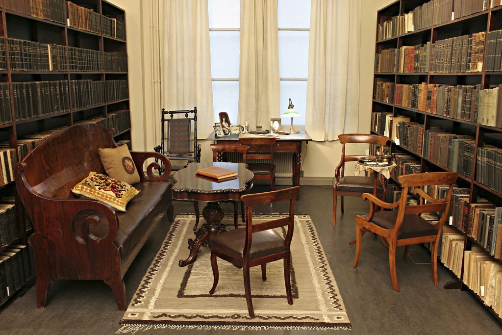 A study turned into a museum: a sofa and coffee table in the foreground, a desk by a window at the back, and a smoking table on the side. The walls are lined with shelves full of books. 