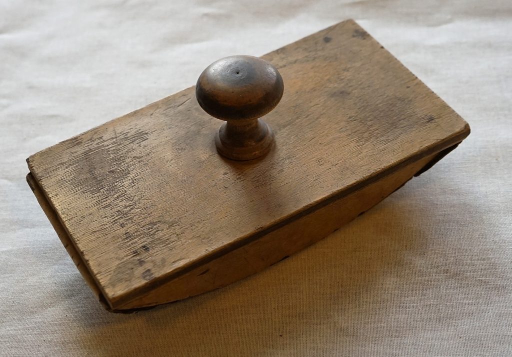 A wooden ink blotter. The round handle is secured to a flat lid. Underneath the lid is a curved base to which blotting paper has been attached.