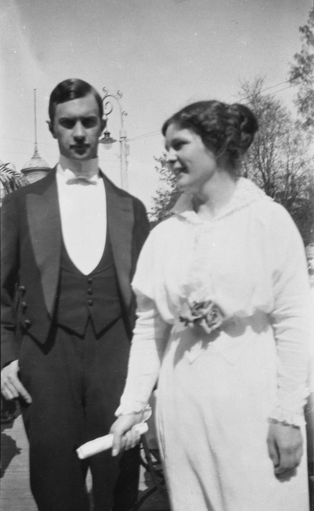 A cropped black-and-white photo taken outdoors on a sunny day showing two people from the knees up standing side by side. On the left is a man in evening dress looking straight at the camera, while on the right is a woman, smiling and relaxed, wearing a white long-sleeve dress, not posing but looking to the left of the camera, as if she were talking to someone.
