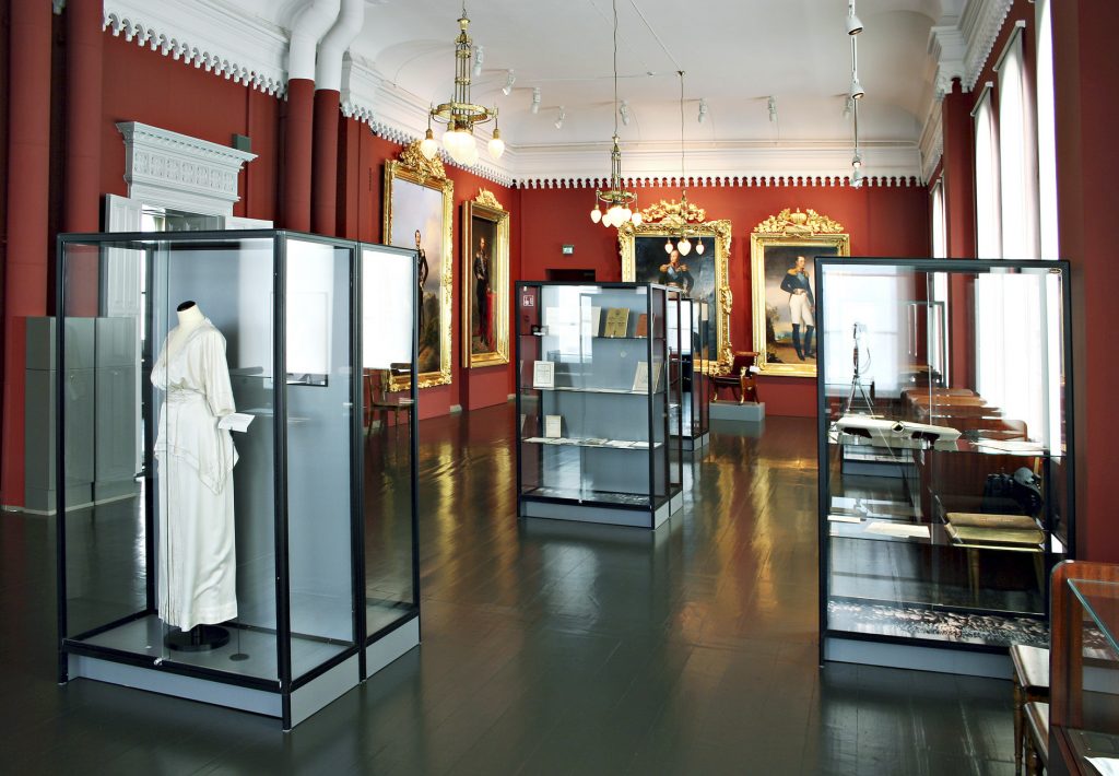 A room with red walls, a white ceiling and a grey floor. On the walls are large gold-framed portraits of Russian emperors, and on the floor are large glass display cases with various objects inside. 