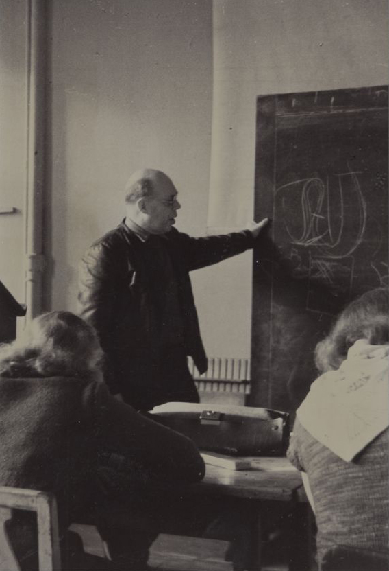 A man is standing at a blackboard, teaching drawing. Two women students are seated at the front with their backs to the camera.
